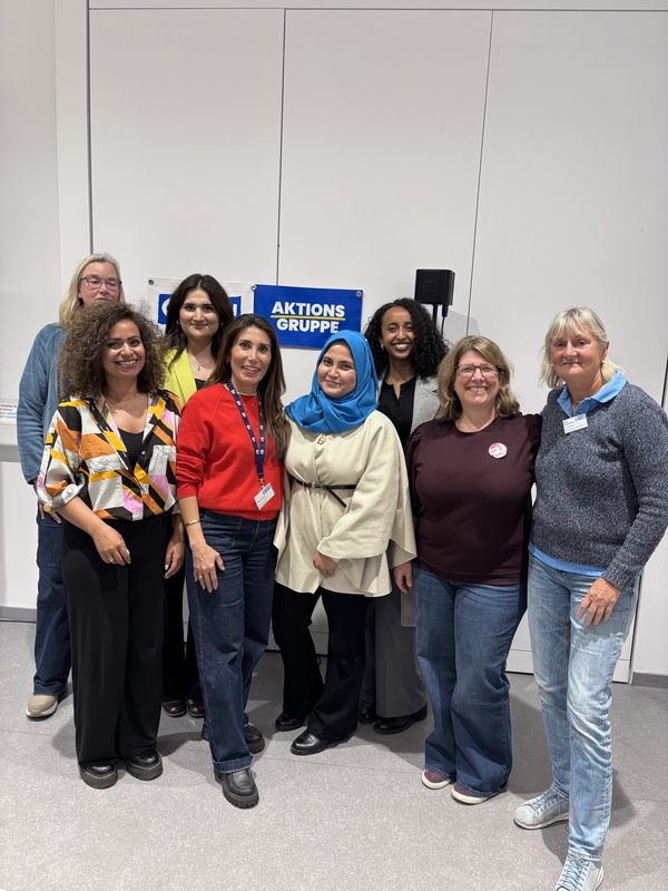 Acht Frauen stehen in einer Reihe vor einer weißen Wand mit der Aufschrift AKTIONS GRUPPE. Sie tragen unterschiedliche legere Business-Kleidung, von Pullovern und Blusen bis hin zu einem blauen Kopftuch, und lächeln gemeinsam in einem hell beleuchteten Innenraum.