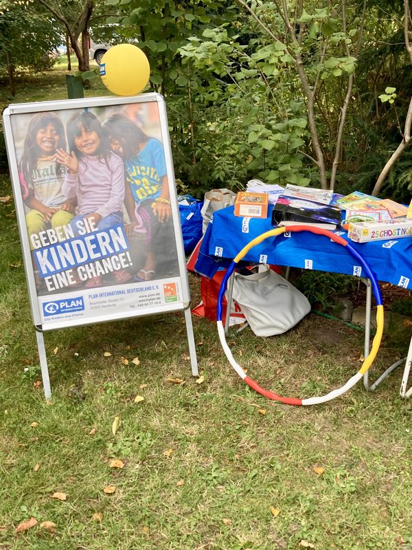 Auf einem Werbeschild von Plan International sind drei lächelnde Kinder zu sehen, darüber steht der deutsche Text GEBEN SIE KINDERN EINE CHANCE!. Daneben stehen auf einem mit einem blauen Tischtuch bedeckten Klapptisch Spielekisten, ein Hula-Hoop-Reifen und eine weiße Tasche auf dem Rasen.