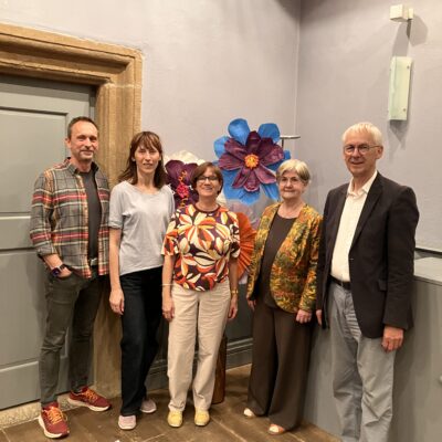 Fünf Erwachsene stehen in einem Raum auf einem Holzboden vor einer Wand, die mit großen, bunten Papierblumen geschmückt ist. Sie lächeln und sind leger gekleidet, bis auf einen Mann im Anzug. Der Raum hat blasslavendelfarbene Wände und eine Tür mit einem Steinrahmen.