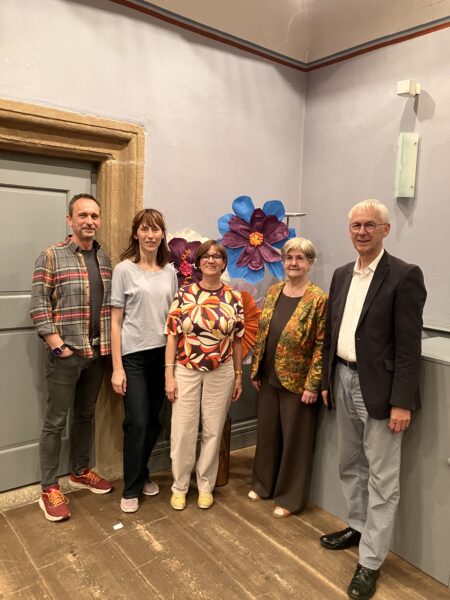 Fünf Erwachsene stehen in einem Raum auf einem Holzboden vor einer Wand, die mit großen, bunten Papierblumen geschmückt ist. Sie lächeln und sind leger gekleidet, bis auf einen Mann im Anzug. Der Raum hat blasslavendelfarbene Wände und eine Tür mit einem Steinrahmen.