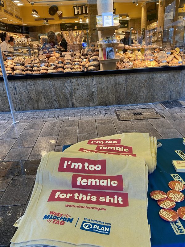 Hellgelbe Tragetaschen mit der Aufschrift Im too female for this shit sind auf einem Tisch vor einer Bäckerei gestapelt. Daneben liegen runde rote Anstecker mit ähnlichen Slogans, während hinter dem Tisch in einer gut gefüllten Bäckereivitrine Reihen von Brot zu sehen sind.