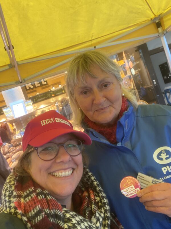 Zwei Frauen stehen unter dem gelben Vordach eines Marktstandes und lächeln. Die Frau im Vordergrund trägt eine rote I am a Girl-Kappe und einen karierten Schal, während die Frau hinter ihr eine blaue Jacke mit einem Anstecker trägt, auf dem Lauf für Mädchen steht.