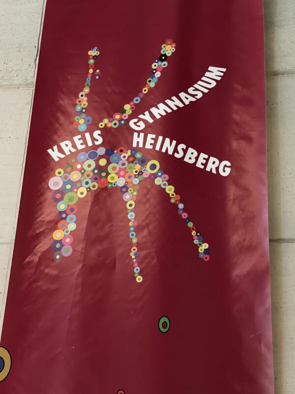 Auf einem vertikalen roten Banner steht in weißer Schrift KREIS GYMNASIUM HEINSBERG. In der Mitte ist eine stilisierte menschliche Figur zu sehen, die aus kleinen, bunten Kreisen besteht und vor einer Wand aus weißen Fliesen positioniert ist.
