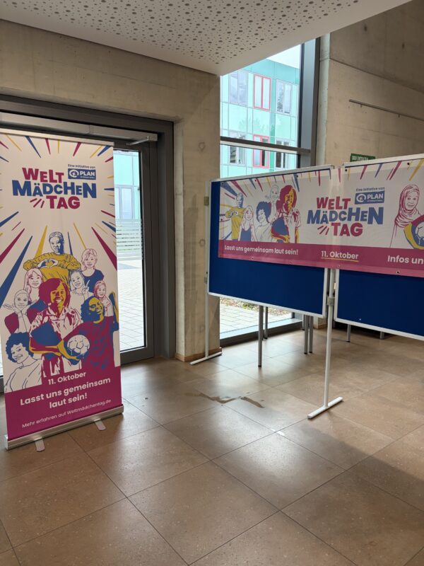 In einem modernen, gefliesten Flur stehen zwei Bannerständer zum Welt-Mädchentag (Internationaler Tag der Mädchen). Darauf sind farbenfrohe Illustrationen verschiedener junger Mädchen und Frauen, das Logo von Plan International sowie das Datum 11. Oktober zu sehen.