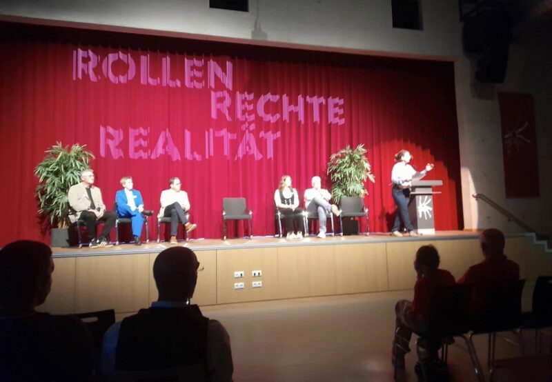 Fünf Personen sitzen auf einer Bühne vor einem roten Vorhang, auf dem der Schriftzug ROLLEN RECHTE REALITÄT zu lesen ist. Eine sechste Person steht an einem Rednerpult und spricht zu den Zuschauern, die sich in einem schwach beleuchteten Saal versammelt haben.