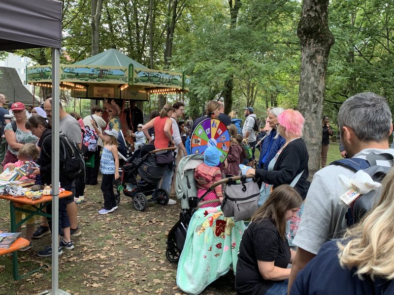 In einem bewaldeten Park steht ein Karussell mit einem bemalten Baldachin, während Familien an einem Bücherstand stöbern. Kinder und Erwachsene stehen auf dem mit Laub bedeckten Boden, und neben einem Kinderwagen, der mit einer gemusterten hellblauen Decke zugedeckt ist, steht ein buntes Glücksrad.
