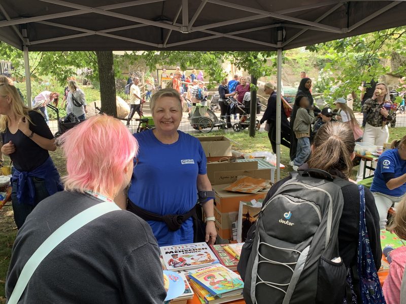 Eine Frau in einem blauen Hemd lächelt hinter einem Tisch, der mit Kinderbüchern bedeckt ist und unter einem Vordach steht. Zu ihrer Linken steht eine Person mit leuchtend pinkfarbenen Haaren, während ihr gegenüber eine weitere Person mit einem schwarzen Rucksack in einer Parkkulisse steht, in der sich viele andere Menschen aufhalten.