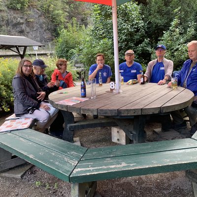 Sechs Personen sitzen draußen um einen großen, runden Picknicktisch aus Holz herum. Sie sind leger in Jacken und T-Shirts gekleidet, haben Getränke vor sich stehen und haben sich unter einem Sonnenschirm in einer grünen Parklandschaft neben einer felsigen Klippe versammelt.