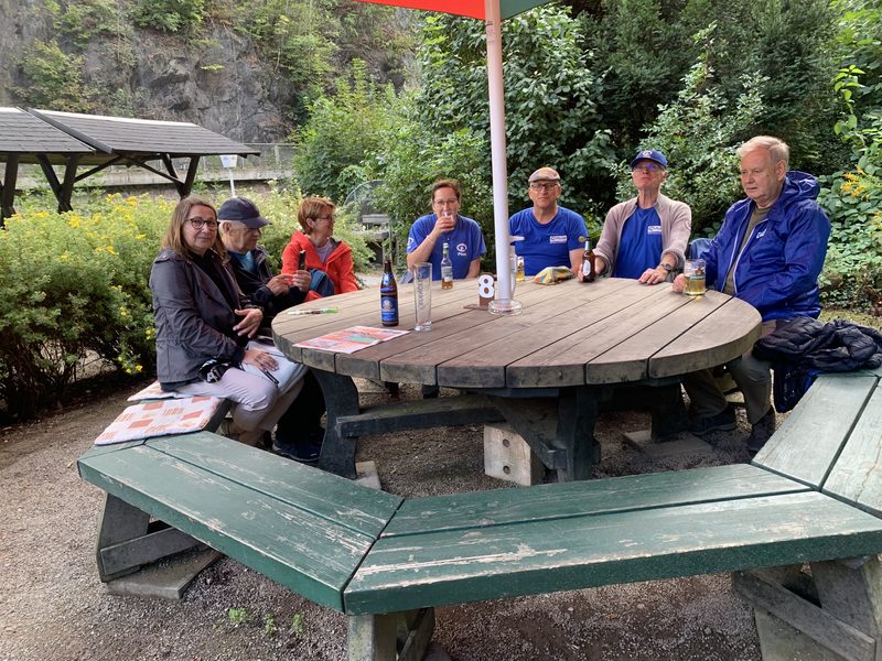 Sechs Personen sitzen draußen um einen großen, runden Picknicktisch aus Holz herum. Sie sind leger in Jacken und T-Shirts gekleidet, haben Getränke vor sich stehen und haben sich unter einem Sonnenschirm in einer grünen Parklandschaft neben einer felsigen Klippe versammelt.