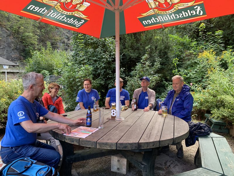 Fünf Personen sitzen im Freien unter einem großen rot-grünen Feldschlösschen-Sonnenschirm um einen runden hölzernen Picknicktisch herum. Sie tragen Freizeitkleidung, darunter mehrere blaue Hemden, und auf dem Tisch stehen Getränke inmitten einer üppig grünen Umgebung.