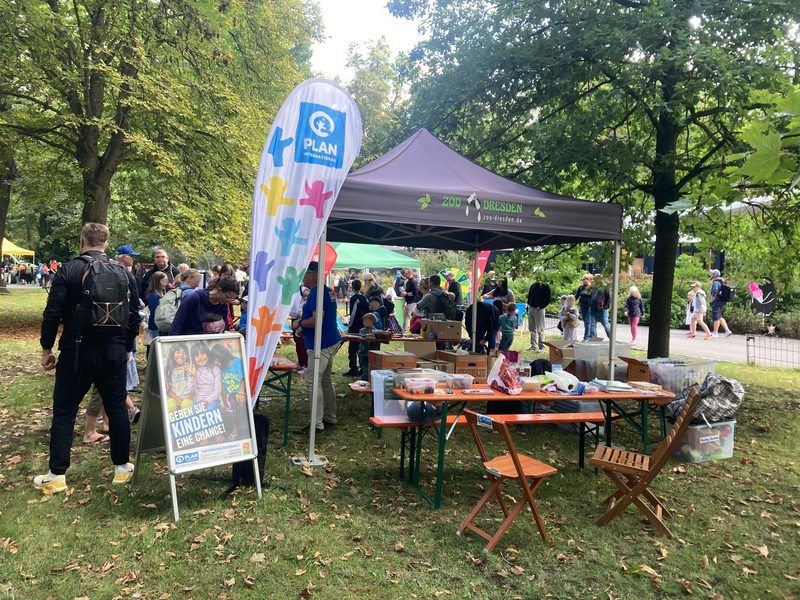 Eine Parkszene zeigt ein Informationszelt des Dresdner Zoos mit Tischen, Stühlen und einer Flagge von Plan International. Menschen schauen sich am Stand um, während ein Schild im Vordergrund ein Foto von zwei lächelnden Kindern mit dem Text Geben Sie Kindern eine Chance zeigt.