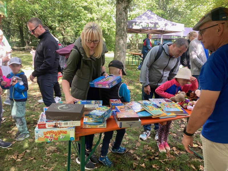 Eine Frau und ein Kind betrachten Brettspiele, die auf einem langen Tisch im Freien ausgebreitet sind. Ein Mann in einem blauen Hemd beobachtet, wie andere Besucher die Artikel in einem sonnendurchfluteten Park mit Rasenfläche durchstöbern. Auf der Tischoberfläche sind mehrere Spiele in Schachteln und Bücher angeordnet.