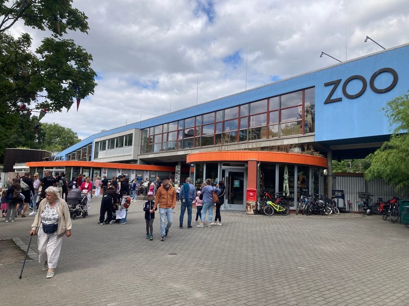Menschen spazieren über einen gepflasterten Platz vor einem blauen Gebäude, auf dessen Dach das Wort ZOO zu lesen ist. Eine Frau im Vordergrund stützt sich auf einen Gehstock, während sich andere, darunter Familien mit Kinderwagen und abgestellten Fahrrädern, am orangefarbenen Eingang versammeln.