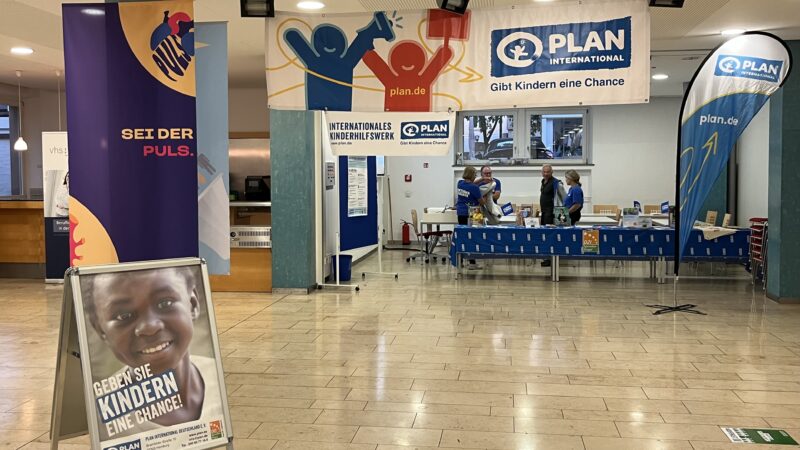 Ein Informationsstand von Plan International in einer Lobby, hinter einem blauen Tisch mit dem Logo der Organisation stehen drei Mitarbeiter. Im Vordergrund ist ein tragbares Schild zu sehen, auf dem das Foto eines lächelnden Kindes und der deutsche Text Geben Sie Kindern eine Chance zu sehen sind.
