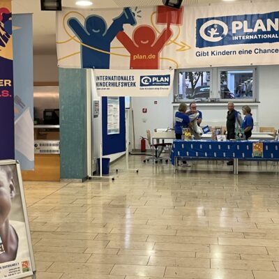 In einer Lobby steht ein Informationsstand von Plan International mit Bannern und einem Schild, auf dem das Porträt eines Kindes zu sehen ist. Hinter einem blauen Tisch stehen drei Mitarbeiter in blauen Hemden bereit, um die Besucher zu betreuen.