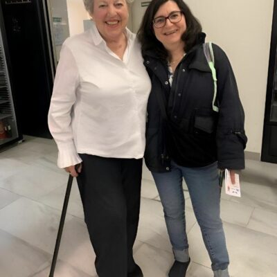 Zwei Frauen lächeln für ein Foto in einem Raum. Links steht eine Frau mit kurzen grauen Haaren, die ein weißes Hemd und eine schwarze Hose trägt und einen Gehstock in der Hand hält. Rechts steht eine Frau mit dunklen Haaren und Brille, die eine marineblaue Jacke, blaue Jeans und eine Umhängetasche trägt.