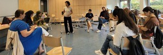 Eine Gruppe von neun Frauen unterschiedlicher Herkunft sitzt im Kreis auf Stühlen in einem Klassenzimmer, während eine Person steht und spricht. Sie tragen Freizeitkleidung und scheinen an einer Besprechung oder einem Workshop teilzunehmen, der in einem Raum mit hellen Holzschränken stattfindet.