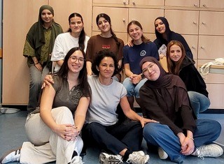 Eine Gruppe von zehn jungen Frauen posiert vor Holzschränken für ein Foto. Sie tragen Freizeitkleidung wie T-Shirts, Jeans und Hijabs und stehen oder sitzen in unterschiedlichen Posen.