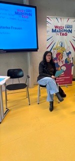 Eine Frau mit dunklen Haaren und Brille sitzt auf einem Stuhl vor einer Präsentationsleinwand und einem Banner zum Welt-Mädchentag. Sie trägt eine dunkle Jacke, blaue Jeans und Stiefel und hält eine blaue Mappe in der Hand, während sie in einen gelben Raum blickt, in dem sich das Publikum befindet.