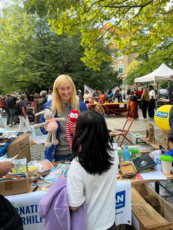 Eine lächelnde Frau steht an einem Marktstand im Freien und hält ein gehäkeltes Kaninchen und ein rot-weiß gestreiftes Affen-Spielzeug in den Händen. Sie steht einer Person mit langen dunklen Haaren gegenüber, umgeben von Kisten mit Spielzeug und Markttischen unter grünen Bäumen.