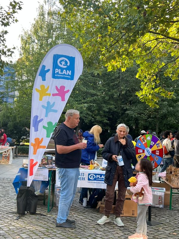 Ein Mann und eine Frau in Outdoor-Kleidung stehen an einem Stand von Plan International in einem Park. Neben einem Tisch steht ein hohes, weißes Banner in Tropfenform mit bunten Figuren im Papierschnitt-Stil, während ein Kind in einer rosa Jacke in der Nähe ein Stofftier in der Hand hält.