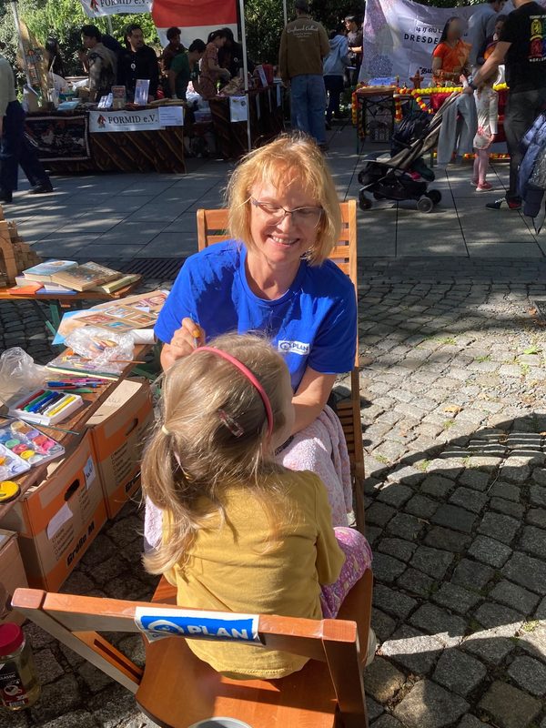 Eine lächelnde Frau in einem blauen Hemd malt einem jungen Mädchen mit blonden Haaren und einem gelben Oberteil das Gesicht an. Sie sitzen auf Holzstühlen auf einem gepflasterten Platz auf einem Markt unter freiem Himmel, im Hintergrund sind verschiedene Stände, Schilder und Menschen zu sehen.