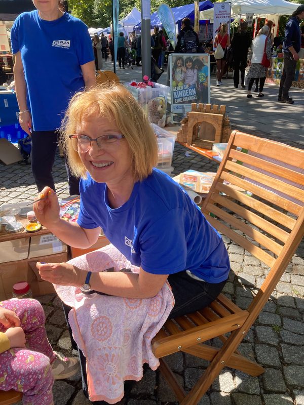 Eine lächelnde blonde Frau in einem blauen T-Shirt und mit Brille sitzt auf einem Holzstuhl, hält einen Pinsel in der Hand und bemalt das Gesicht eines Kindes. Sie befinden sich auf einem Straßenfest im Freien mit Marktständen, bunten Bannern und Menschen, die im Hintergrund vorbeigehen.