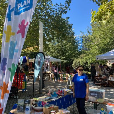 Eine Frau in einem blauen T-Shirt von Plan International steht bei einer Veranstaltung im Freien im Park hinter einem Tisch. Auf dem Tisch liegen verschiedene Gegenstände und Werbematerialien, während auf dem sonnigen, von Bäumen gesäumten Platz große Werbebanner im Wind flattern.