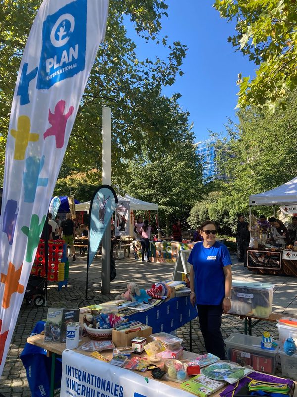 Eine Frau in einem blauen T-Shirt von Plan International steht bei einer Veranstaltung im Freien im Park hinter einem Tisch. Auf dem Tisch liegen verschiedene Gegenstände und Werbematerialien, während auf dem sonnigen, von Bäumen gesäumten Platz große Werbebanner im Wind flattern.
