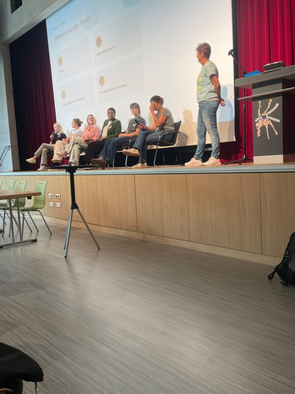 Fünf Diskussionsteilnehmer sitzen in einer Reihe auf einer Bühne vor einer Leinwand, während ein Moderator rechts von ihnen steht. Die Bühne ist mit dunkelroten Vorhängen versehen, und auf dem Podium im Vordergrund ist ein farbenfrohes, kreisförmiges Logo zu sehen.