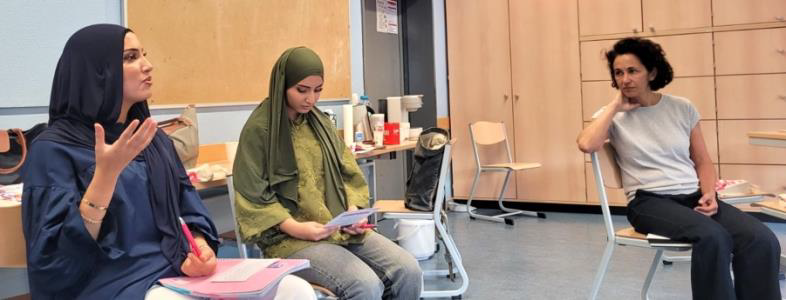 Drei Frauen sitzen in einem Klassenzimmer im Kreis. Zwei von ihnen tragen ein Kopftuch und langärmelige Oberteile, während sie über Unterlagen sprechen; eine dritte Frau mit lockigem Haar sitzt zu ihrer Rechten und hört zu. Der Raum hat hellblaue Wände und Aufbewahrungsschränke aus Holz.
