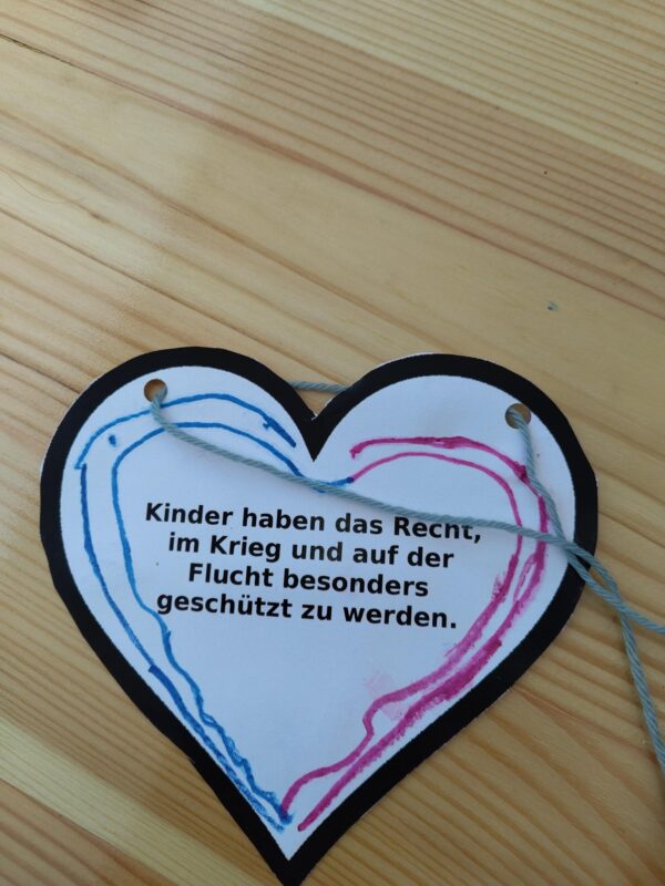 Ein herzförmiger Papierschnitt mit Zeichnungen in Blau und Rosa, die mit Filzstift angefertigt wurden. Darauf steht: Kinder haben das Recht, im Krieg und auf der Flucht besonders geschützt zu werden.
