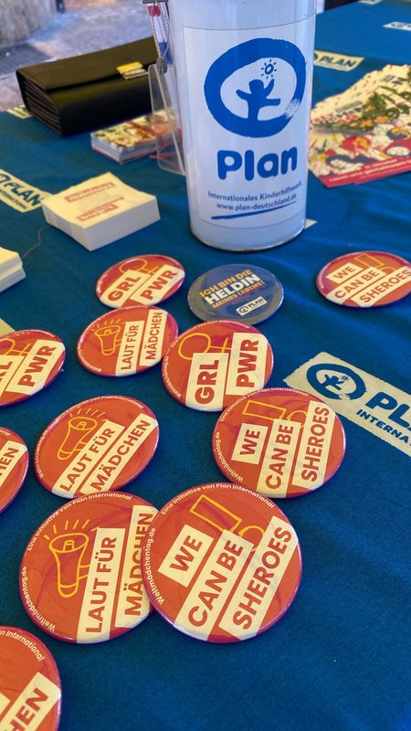 Auf einer blauen Tischdecke liegen verschiedene Werbe-Buttons von Plan International verstreut. Sie sind in Rot und Gelb gestaltet und tragen motivierende Slogans wie GRL PWR, LAUT FÜR MÄDCHEN und WE CAN BE SHEROES auf Englisch und Deutsch.