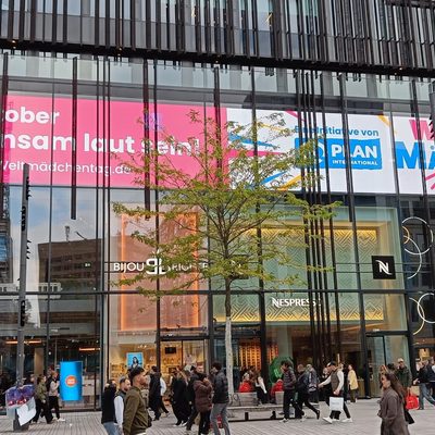An der Glasfassade eines modernen Gebäudes sind bunte Banner zu sehen, die für den Internationalen Mädchentag werben. Im Vordergrund erstreckt sich ein belebter Fußgängerplatz, auf dem sich Menschen unterschiedlichster Herkunft in herbstlicher Kleidung tummeln; dabei sind verschiedene Ladenschilder zu erkennen.