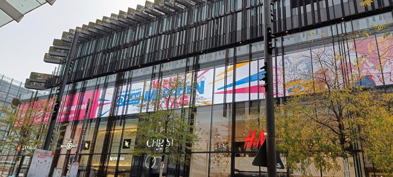 An einem modernen, mehrstöckigen Glasgebäude prangt ein großes, quer verlaufendes Werbebanner für Plan International. Unterhalb des Banners sind durch die Glasfassade die Ladenschilder von H&M und Christ zu sehen, eingerahmt von herbstlichen Bäumen vor einem bewölkten Himmel.