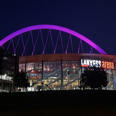 Die Lanxess Arena bei Nacht: Das leuchtend violette, gewölbte Dach hebt sich deutlich vom dunkelblauen Himmel ab. Die moderne Glaskonstruktion wird von innen beleuchtet, und der Name der Arena ist auf der Fassade über einem grasbewachsenen Vordergrund deutlich zu erkennen.