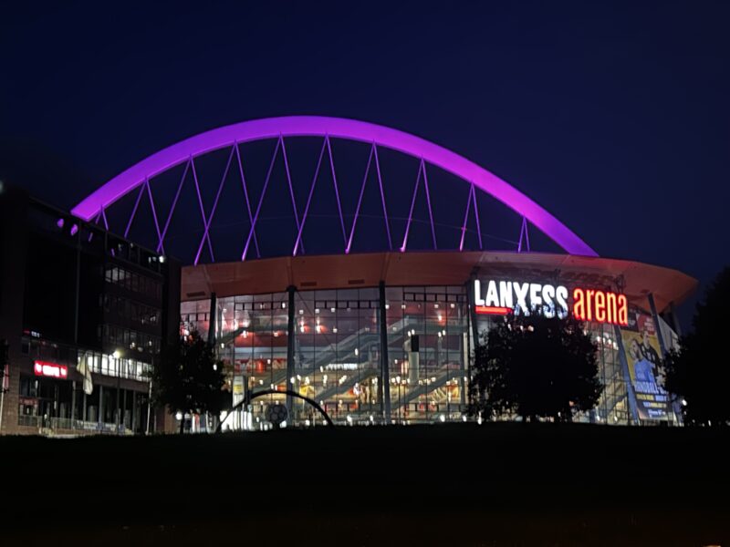 Die Lanxess Arena bei Nacht: Das leuchtend violette, gewölbte Dach hebt sich deutlich vom dunkelblauen Himmel ab. Die moderne Glaskonstruktion wird von innen beleuchtet, und der Name der Arena ist auf der Fassade über einem grasbewachsenen Vordergrund deutlich zu erkennen.